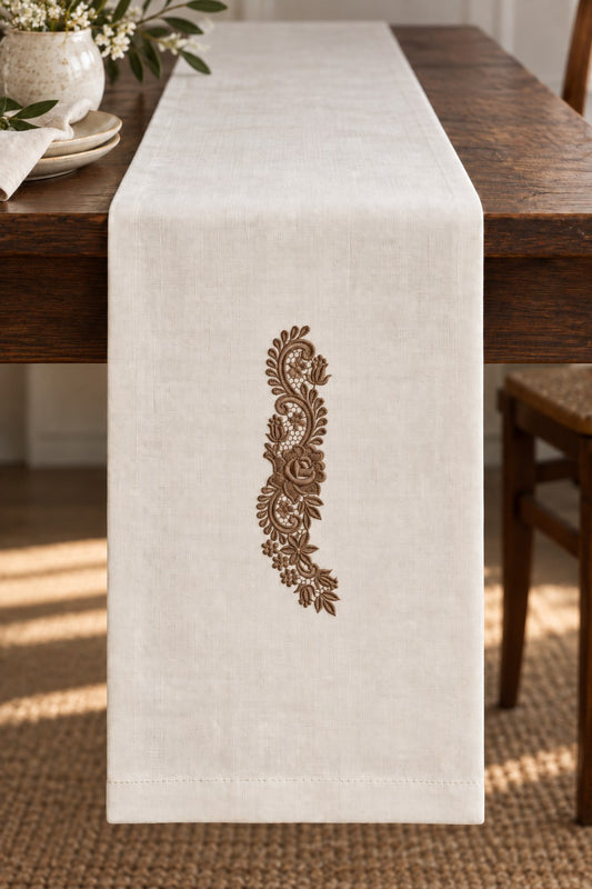 Styled linen table runner mockup with a single muted dark bronze brown rose and tulip lace spray embroidered on pale linen; the complete motif has clear fabric around it.