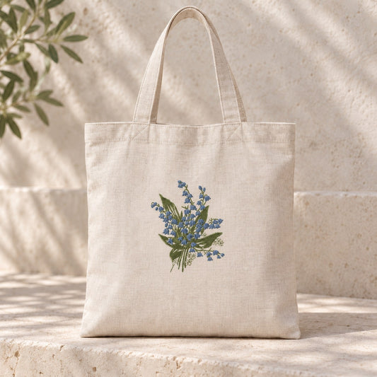Dusty blue lily of the valley flowers and sage leaves embroidered on an oatmeal linen tote resting on a stone bench.