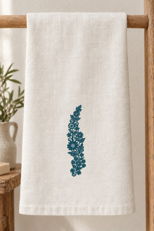 Styled linen hand towel mockup with a single deep muted petrol teal daisy meadow lace vine embroidered on pale linen; the complete motif has clear fabric around it.