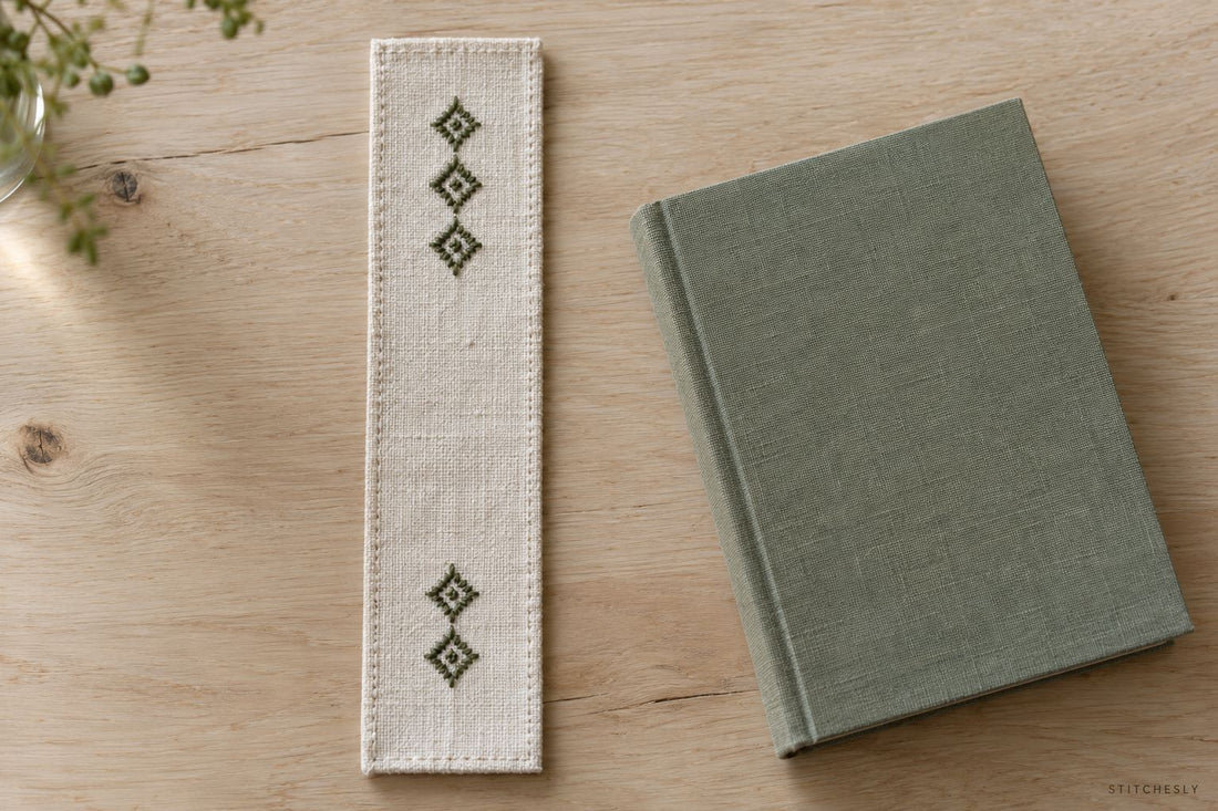 Narrow ivory linen bookmark with two separated groups of sage geometric embroidery beside a closed clothbound book.