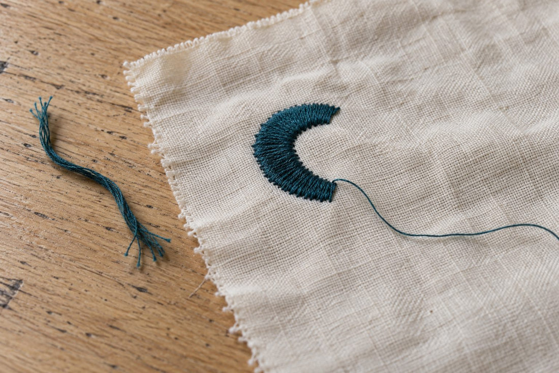 Cream embroidery sample with a teal stitched curve and loose thread ends on a wooden table