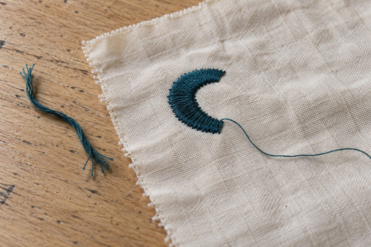 Cream embroidery sample with a teal stitched curve and loose thread ends on a wooden table