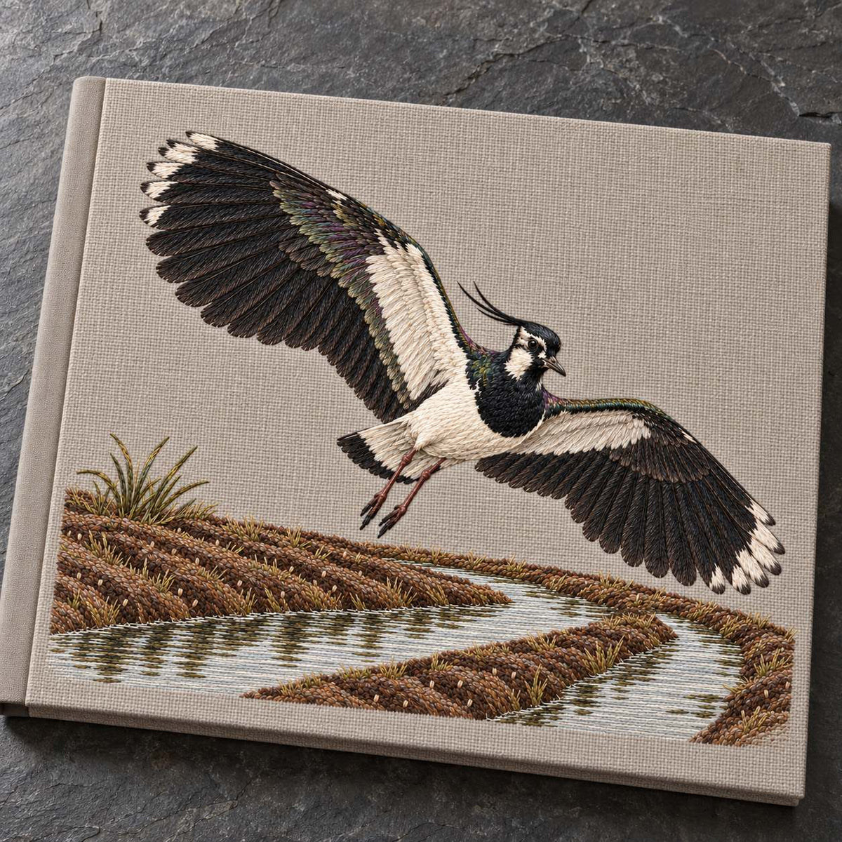 Lapwing Over the Wet Furrow visual mockup shown on a cloth portfolio cover, with the design on warm grey linen.