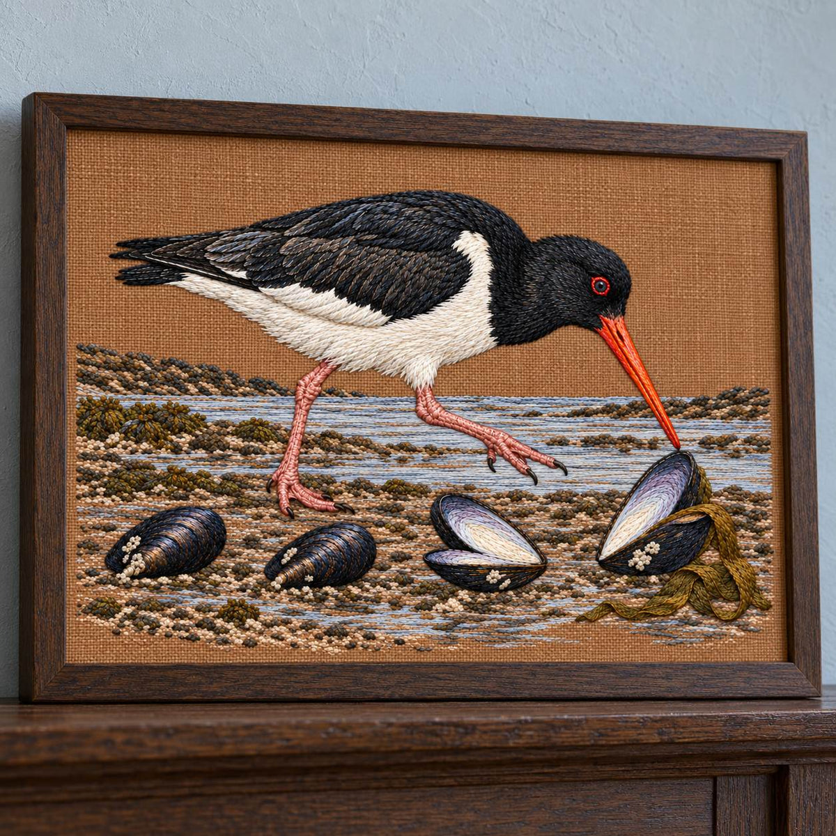 Oystercatcher Across the Mussel Line visual mockup shown in a frame leaning on a mantel, with the design on warm ochre linen.