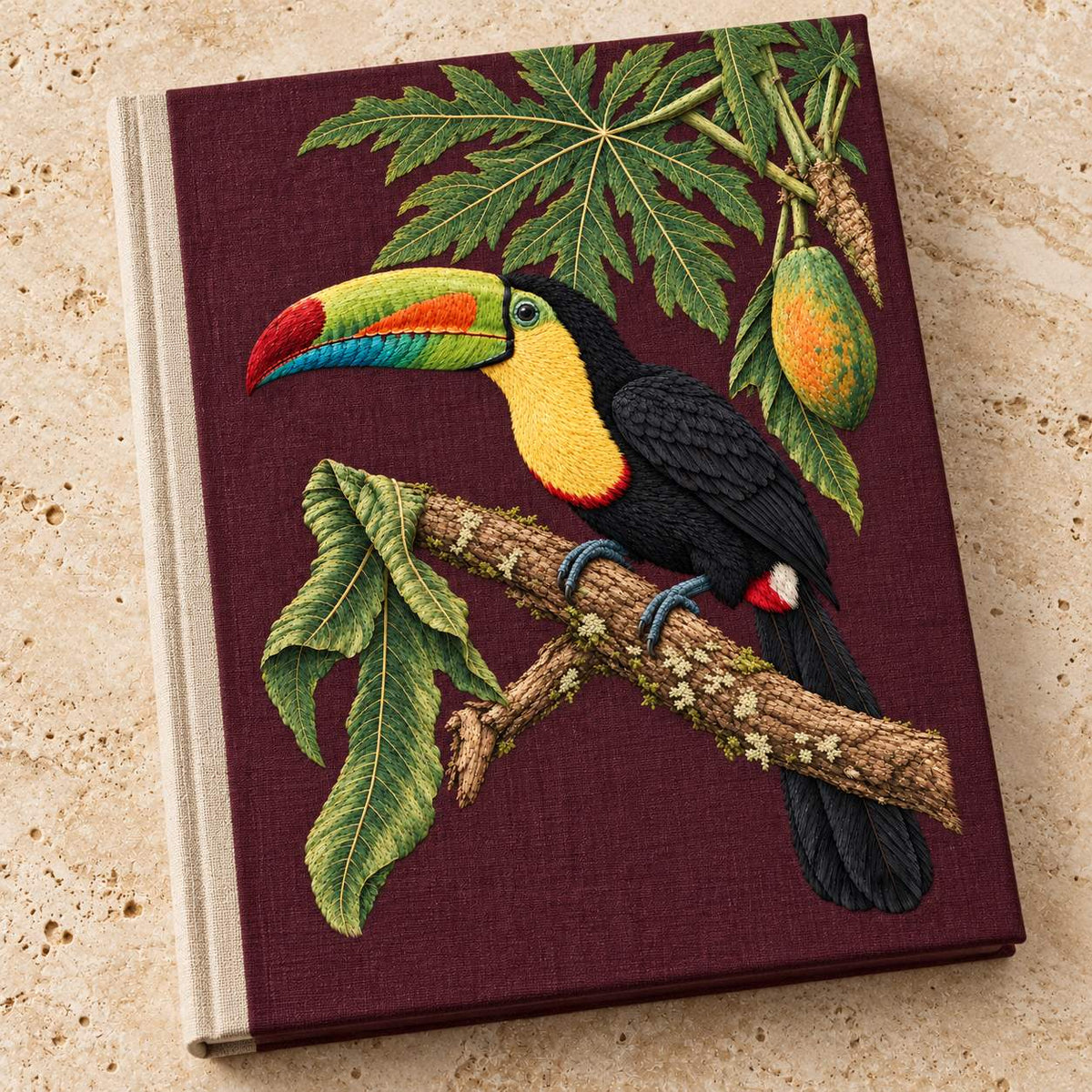 Toucan Under the Papaya Canopy visual mockup shown on a cloth portfolio cover, with the design on deep burgundy linen.