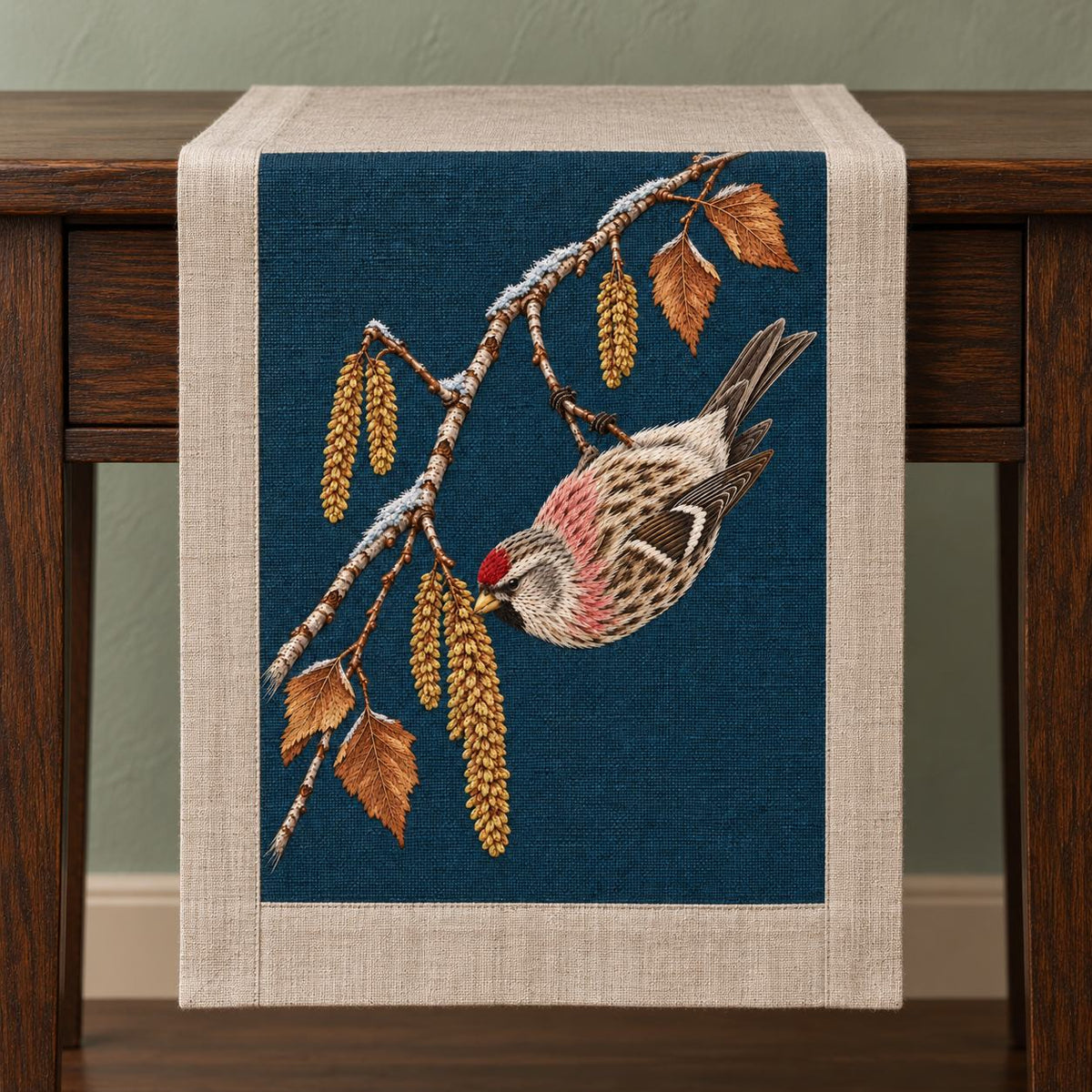 Redpoll Beneath the Birch Snow Tassel visual mockup shown on a textile runner over a console, with the design on dark teal-blue linen.