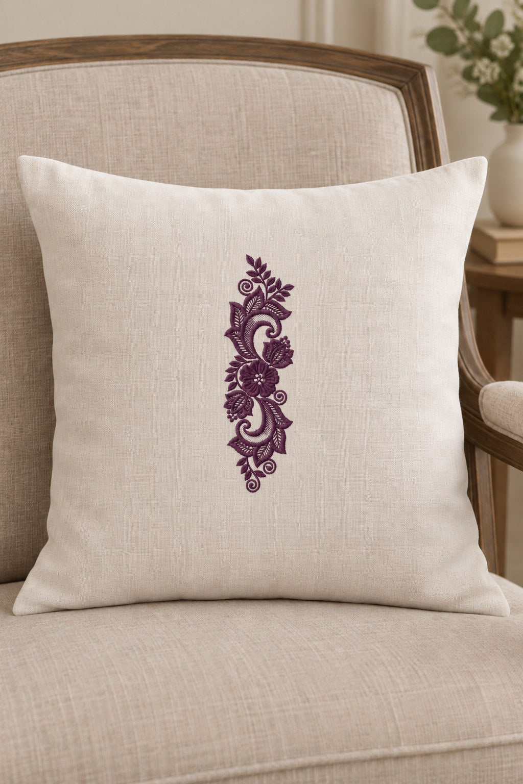 Styled linen cushion mockup with a single deep muted aubergine purple lace scroll floral ornament embroidered on pale linen; the complete motif has clear fabric around it.