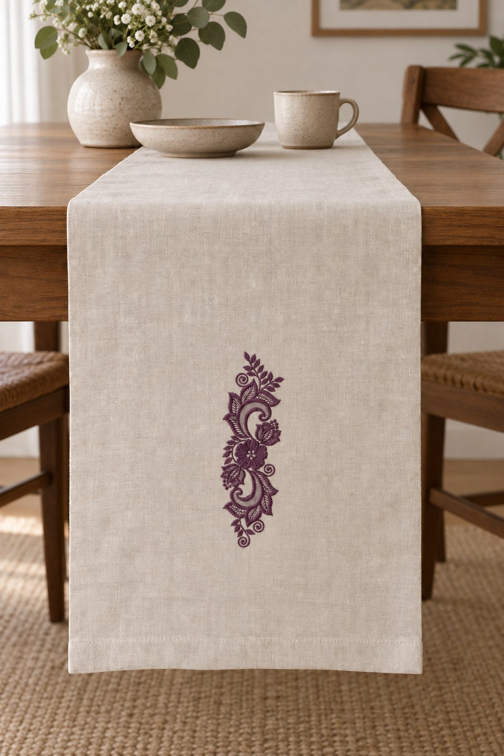 Styled linen table runner mockup with a single deep muted aubergine purple lace scroll floral ornament embroidered on pale linen; the complete motif has clear fabric around it.