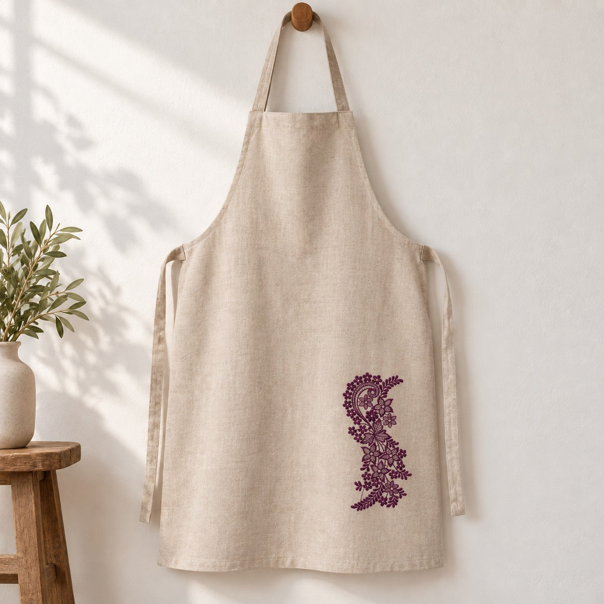 Natural linen apron hanging from a wooden peg with one small aubergine lace-style vine on the lower skirt.