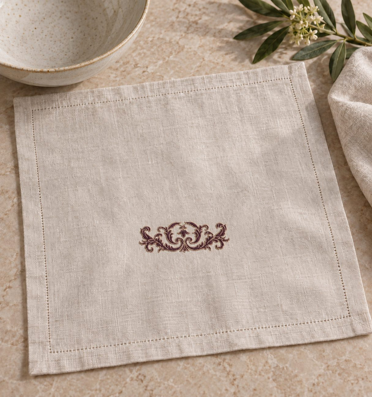 Flax linen dinner napkin with a compact aubergine and parchment crest beside a ceramic bowl.