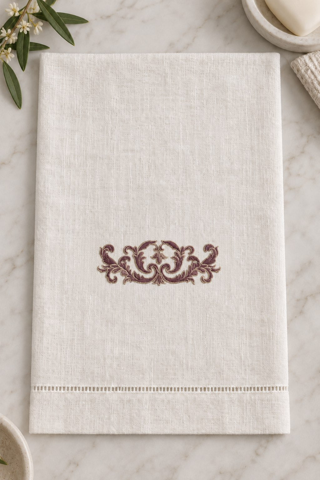 Open ivory guest towel with a centered two-tone symmetrical acanthus ornament above a hemstitched edge.