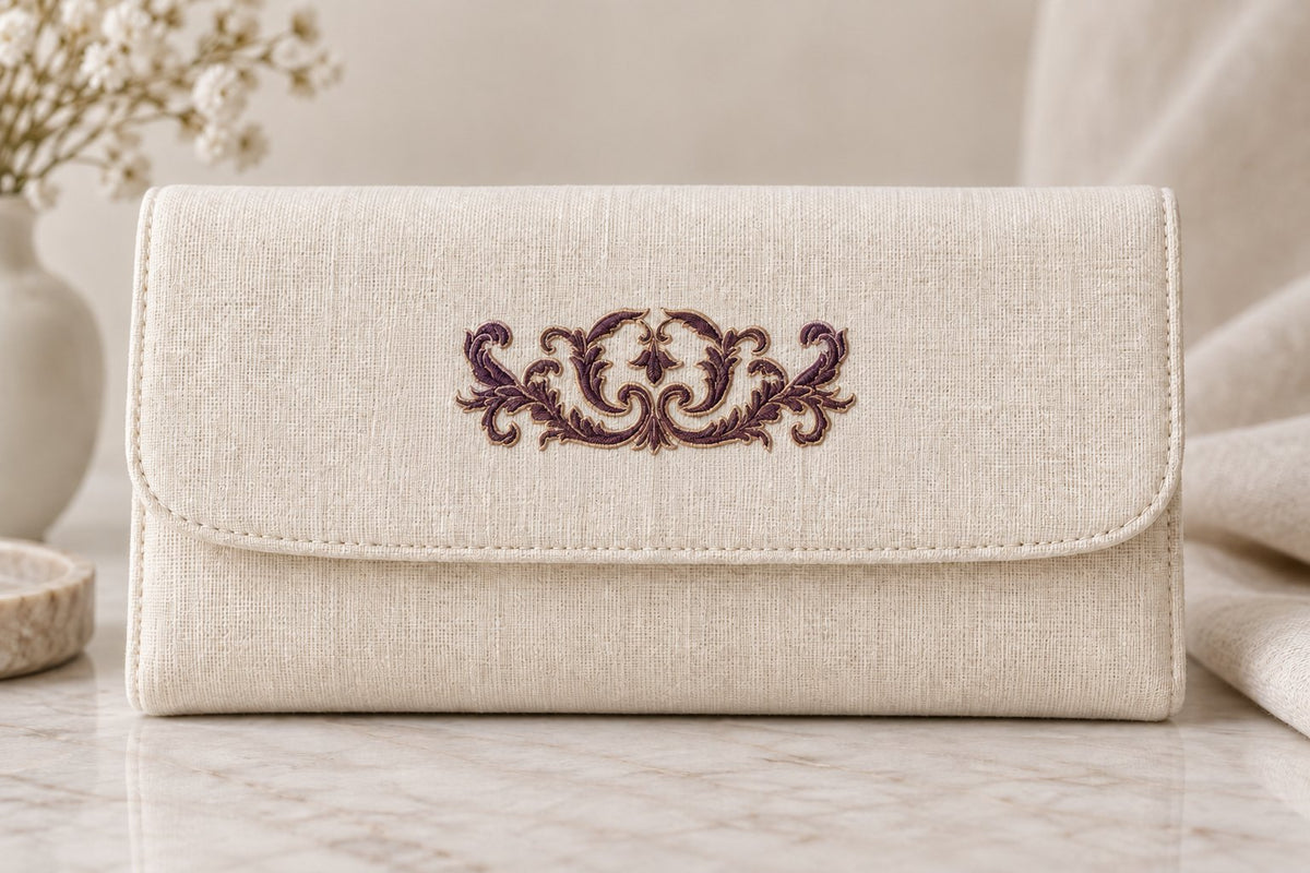 Ivory linen clutch with a small symmetrical aubergine acanthus crest and pale edging on the flap.