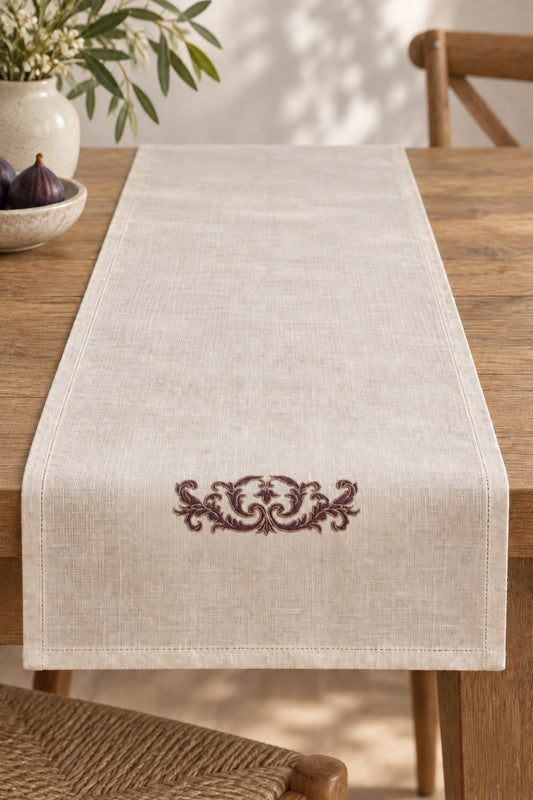 Ivory linen table runner with a small aubergine acanthus crest outlined in parchment near its short end.