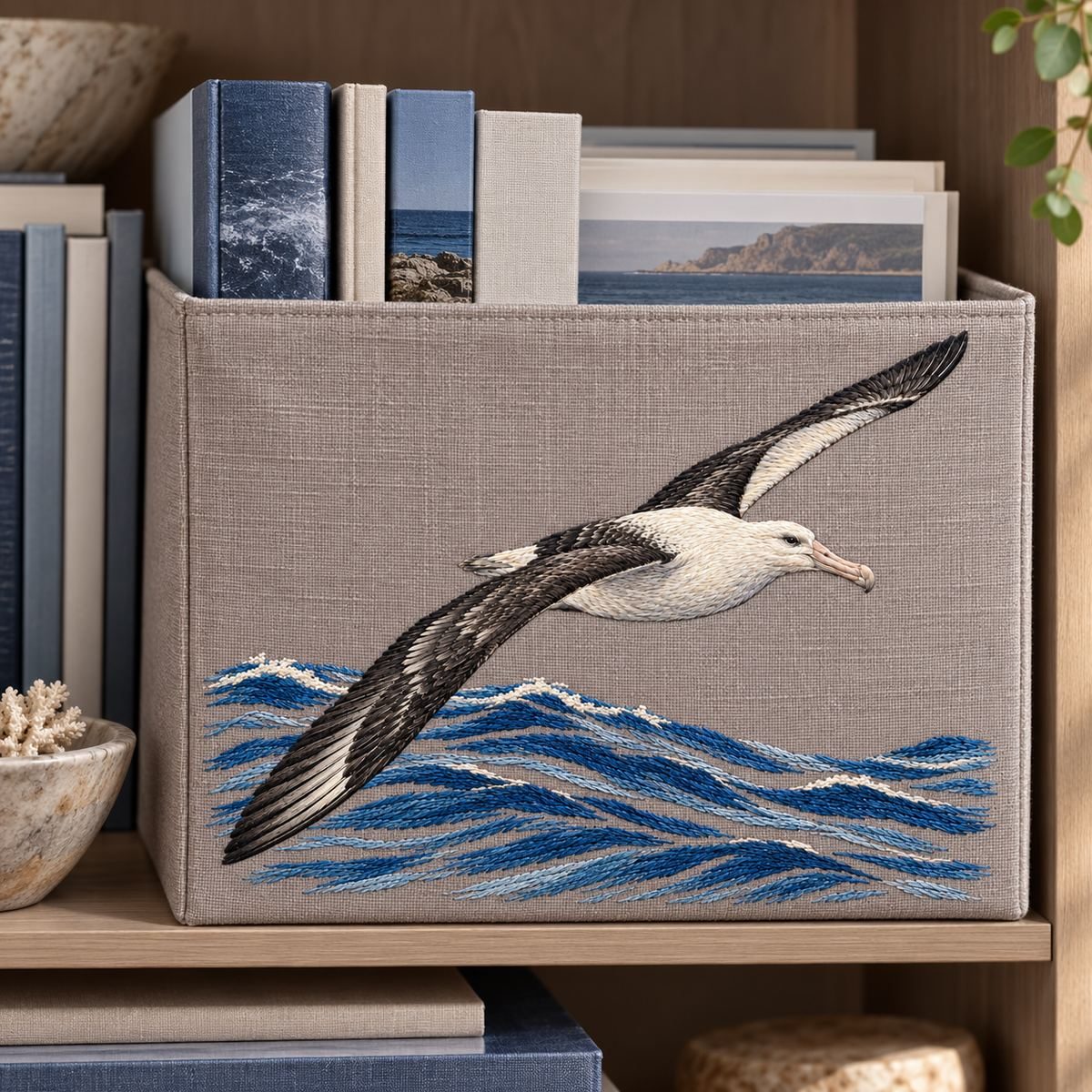 Albatross embroidery design in a digital mockup of wide taupe fabric-covered bookshelf magazine bin front beside marine books. Placement inspiration for Study 03.