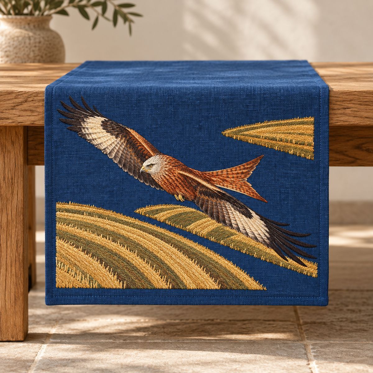 Kite embroidery design in a digital mockup of An indigo rectangular bench-end hanging panel, draping vertically from a long oak bench but the embroidered composition stays LANDSCAPE on its wide upper face. Placement inspiration for Study 05.
