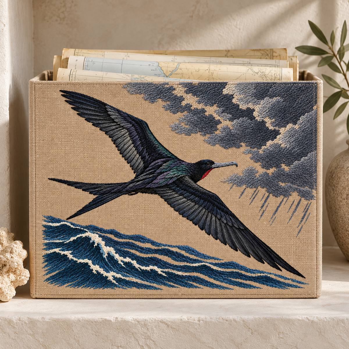 Frigatebird embroidery design in a digital mockup of A mushroom-linen rectangular storage-box front for folded sea charts, standing on a pale plaster niche. Placement inspiration for Study 06.