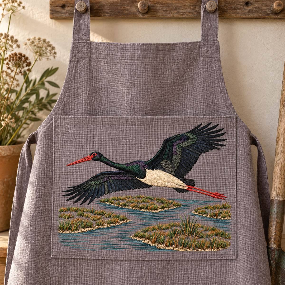 Black Stork embroidery design shown in a digital mockup of A plum-grey linen landscape pocket panel on a large gardener apron hung flat on pegs, pocket wide enough for full marsh design. Study 08.