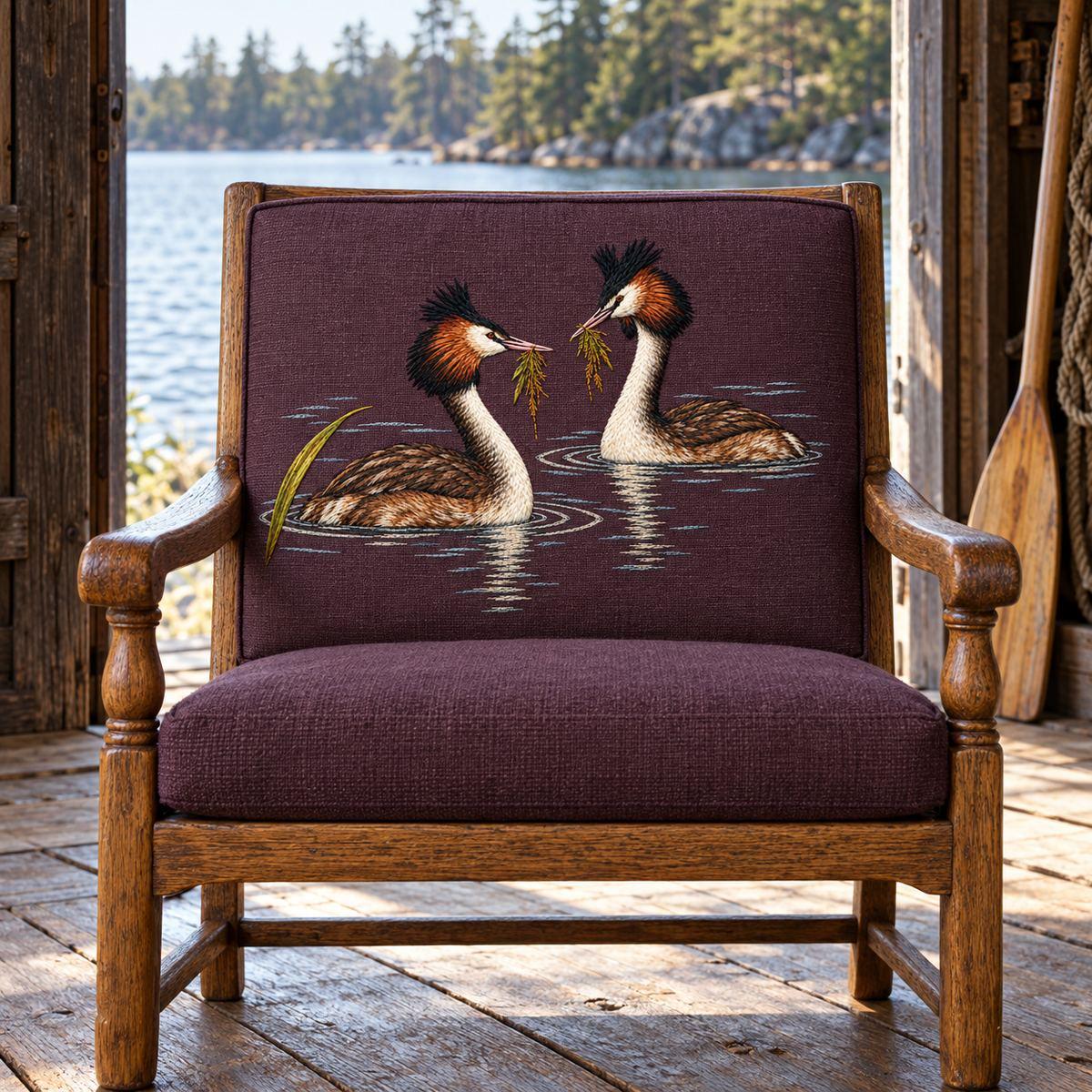 Grebe Pair embroidery design in a digital mockup of A removable embroidered back panel on a small oak lakeside reading chair, viewed straight-on through an open boathouse doorway; soft water light and aged timber, no foreground obstruction. Placement inspiration for Study 15.