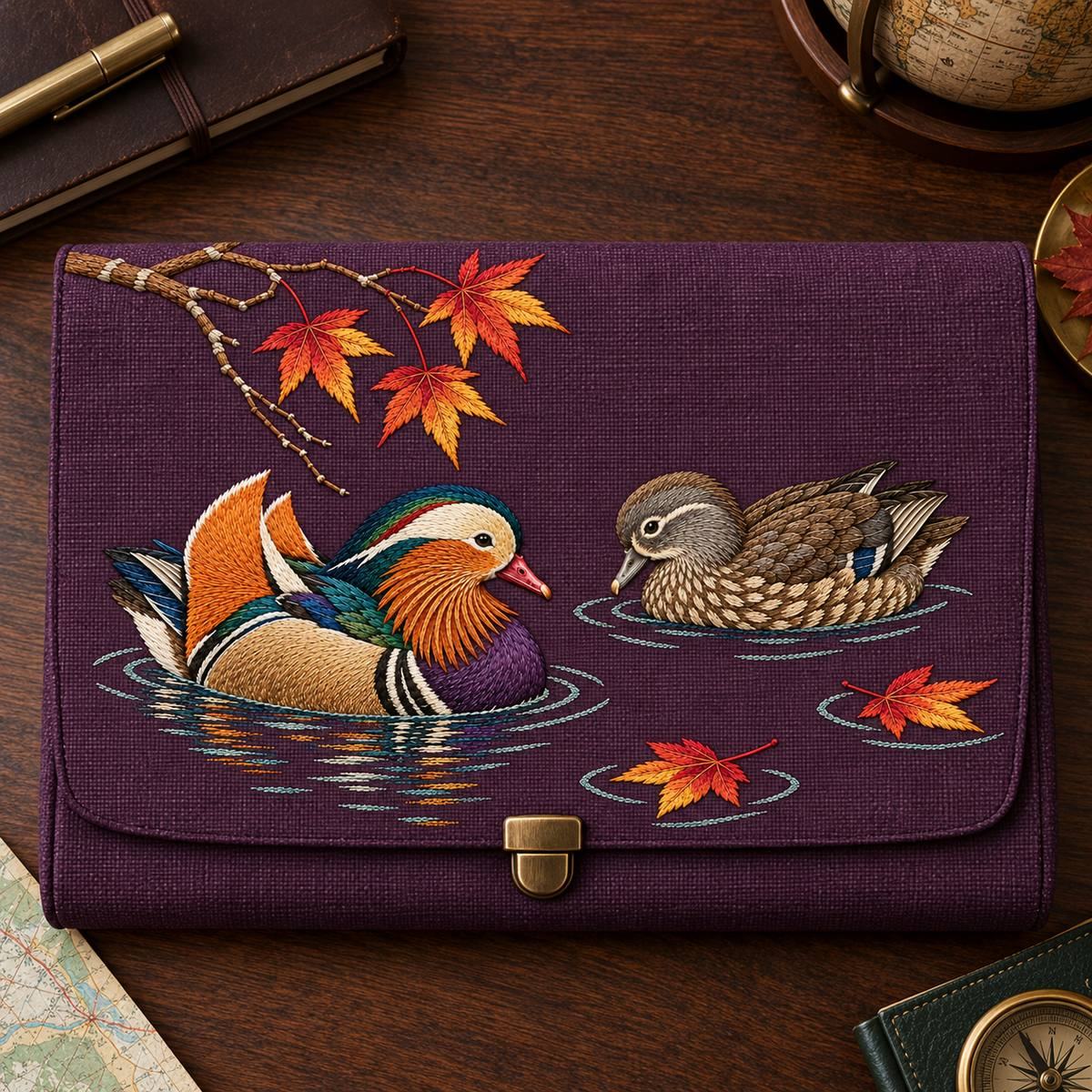 Mandarin Duck Pair embroidery design in a digital mockup of A broad structured travel document clutch on a dark walnut desk, top-down photography, the full landscape embroidery on the front and a small brass clasp outside the design. Placement inspiration for Study 19.