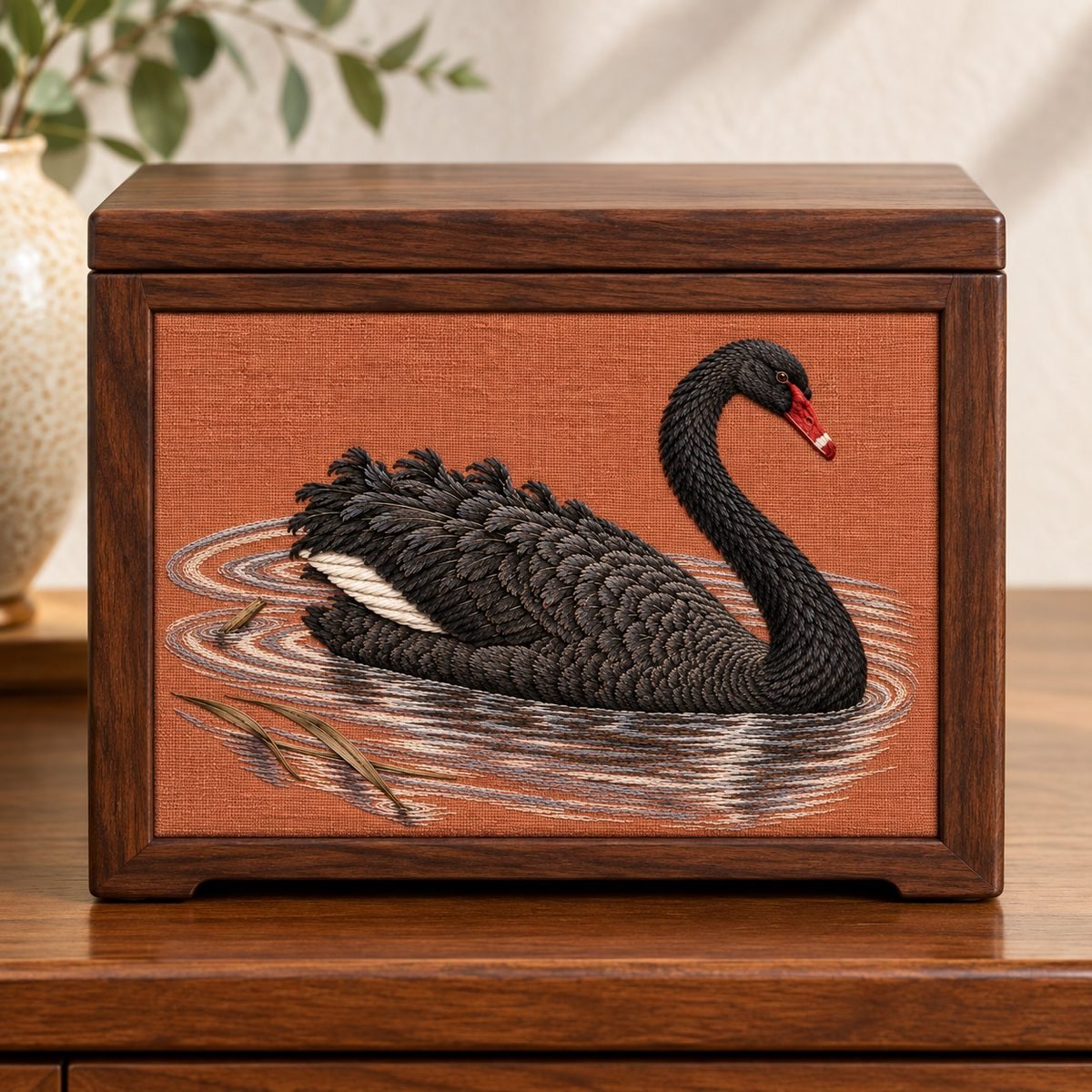 Black Swan embroidery design in a digital mockup of A landscape terracotta embroidery inset in the front of a small portable dark walnut jewellery chest, all handles on sides outside art, chest closed and front viewed squarely. Placement inspiration for Study 32.