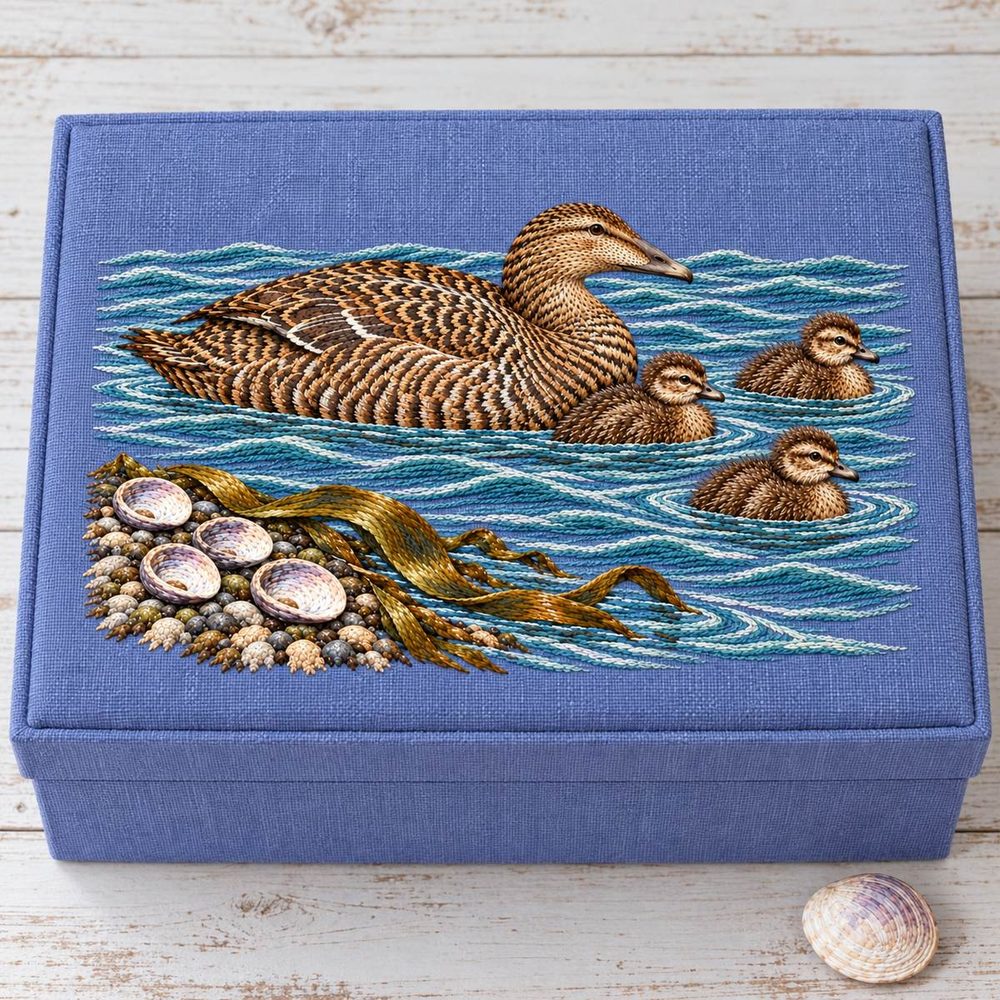 Eider and Chicks embroidery design in a digital mockup of lavender-blue linen nursery memory chest with embroidery inset into flat lid, overhead on whitewashed wood; one plain shell beside it. Placement inspiration for Study 34.