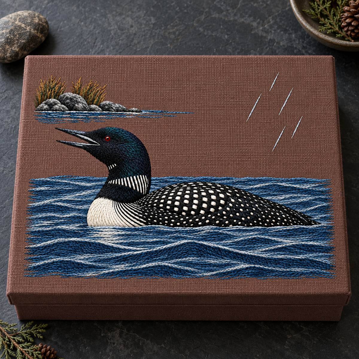 Loon embroidery design in a digital mockup of horizontal plum-brown linen map archive box with flat embroidered lid, overhead on dark slate desk, small stone at side. Placement inspiration for Study 35.