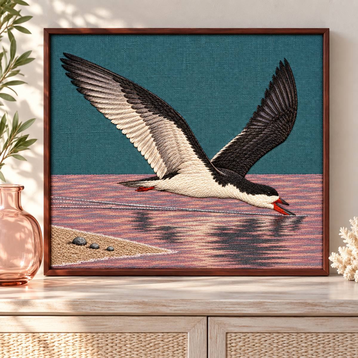 Skimmer embroidery design shown in a digital mockup of teal linen embroidery inset in a slim rosewood frame, front-on above a pale coastal console, one rose glass vessel beside frame. Study 37.