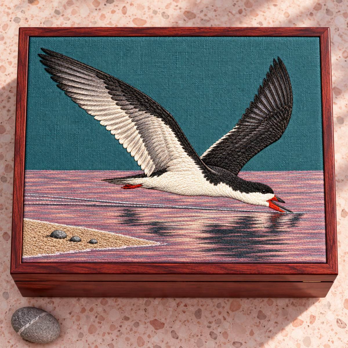 Skimmer embroidery design in a digital mockup of teal linen flat lid of a rose-stained timber keepsake chest, overhead on pale pink terrazzo, single smooth grey pebble outside. Placement inspiration for Study 37.