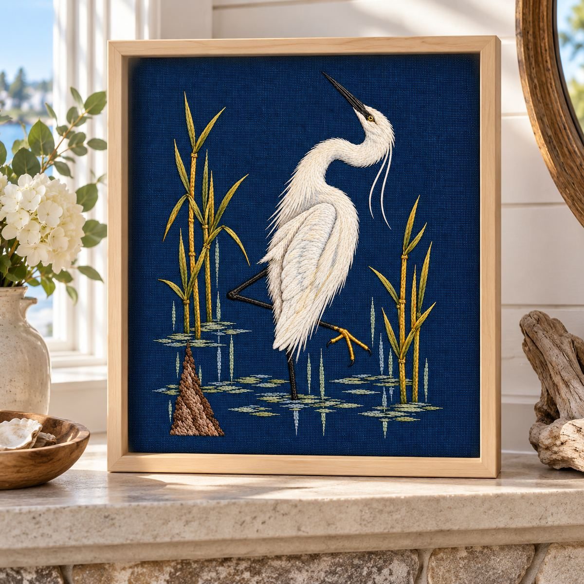 Egret embroidery design in a digital mockup of portrait textile on a pale maple shadowbox standing on a waterside cottage mantel. Placement inspiration for Study 39.