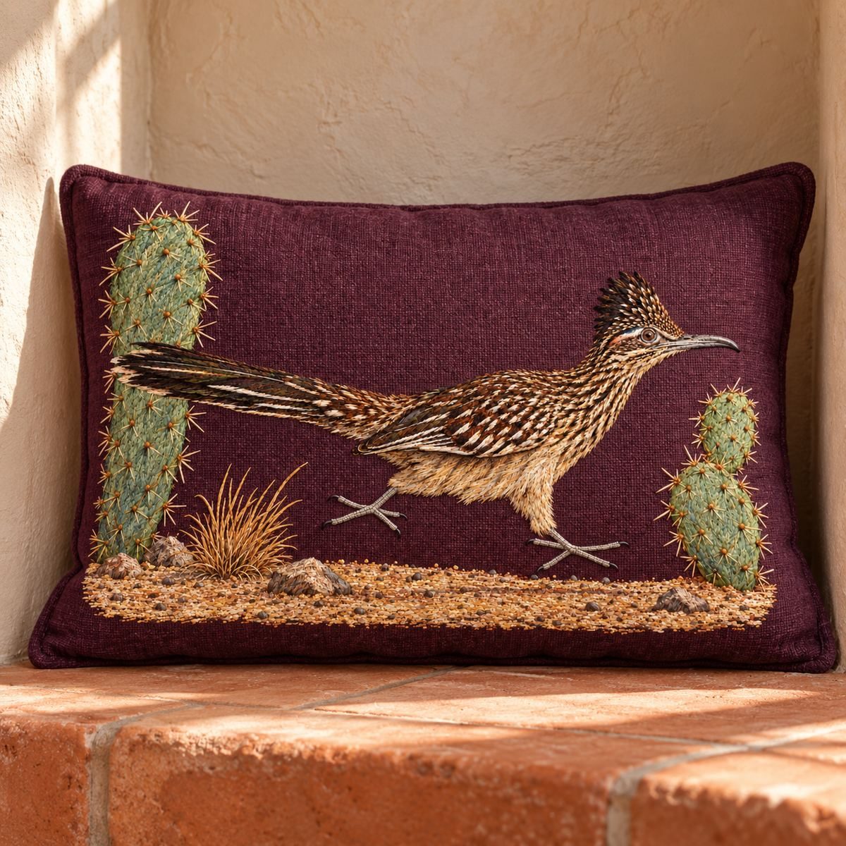 Roadrunner embroidery design in a digital mockup of Wide small lumbar pillow in an adobe window niche with pale plaster and terracotta floor. Complete landscape artwork, no extra cactus decorating the fabric. Placement inspiration for Study 47.