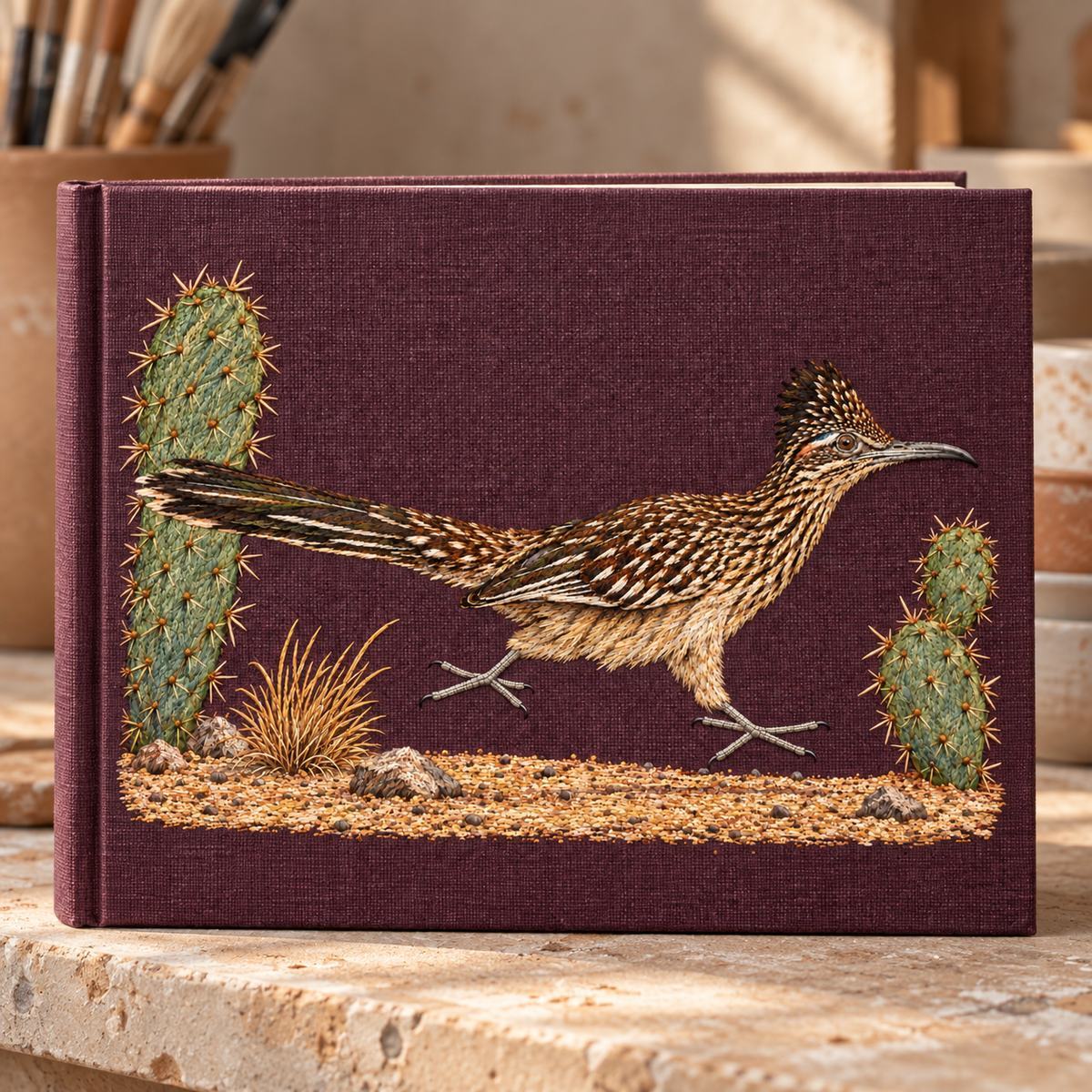 Roadrunner embroidery design in a digital mockup of Landscape linen sketchbook , closed upright on a pottery studio ledge. Plain spine, stitched roadrunner cover, no text or logos. Placement inspiration for Study 47.