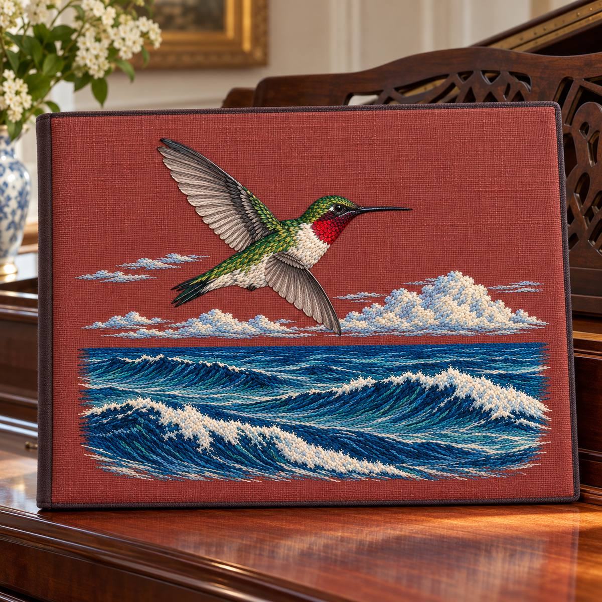 Ruby-throated Hummingbird embroidery design in a digital mockup of A large landscape music manuscript portfolio with rust-plum linen front and plain dark edging, standing on a piano away from keys, no musical text on cover. Placement inspiration for Study 63.