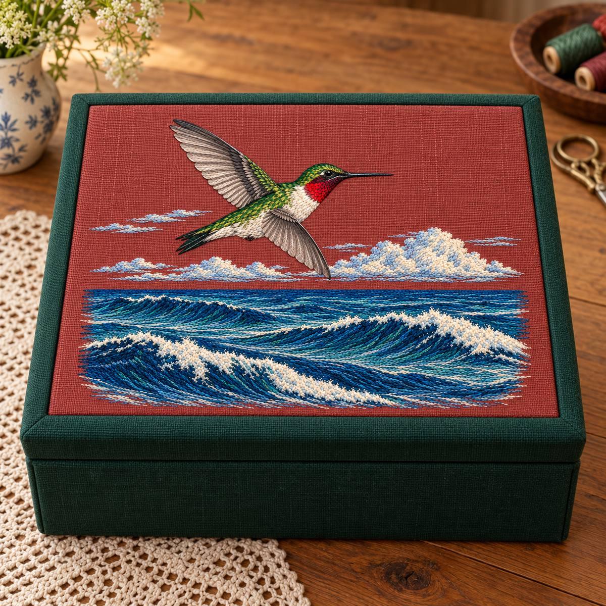 Ruby-throated Hummingbird embroidery design in a digital mockup of A horizontal embroidery inset in the outside lid of a deep green sewing box with rust-plum linen panel, closed on a wooden table, scissors elsewhere. Placement inspiration for Study 63.