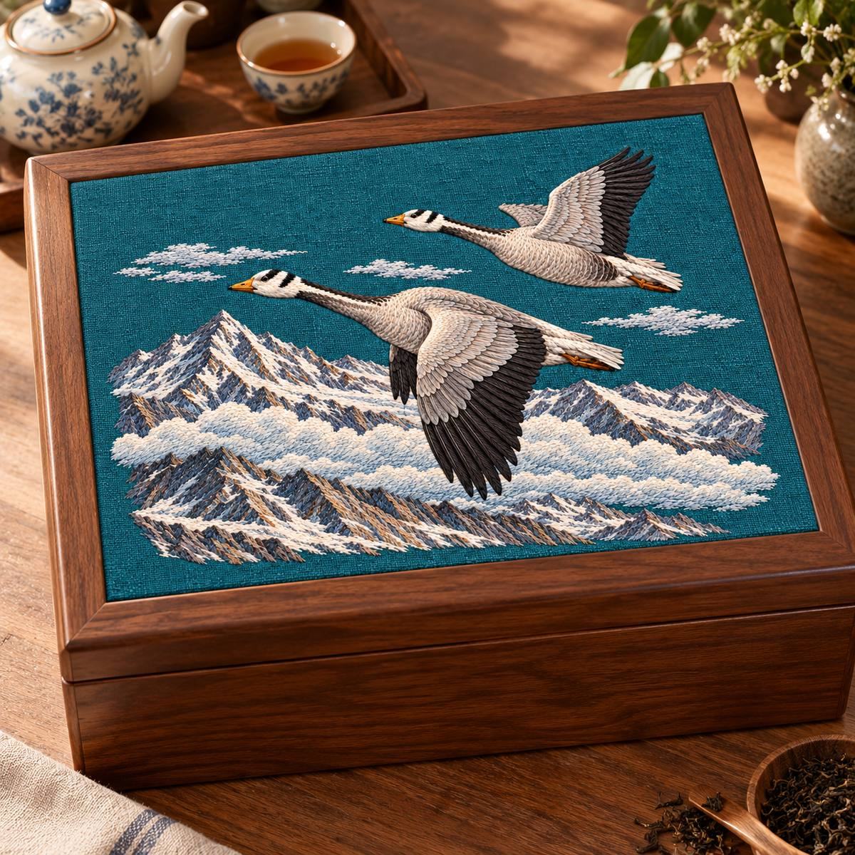 Bar-headed Goose Pair embroidery design in a digital mockup of A horizontal embroidered insert on the closed lid of a large tea service chest, warm walnut and teal linen, angled tabletop view with motif completely readable. Placement inspiration for Study 64.