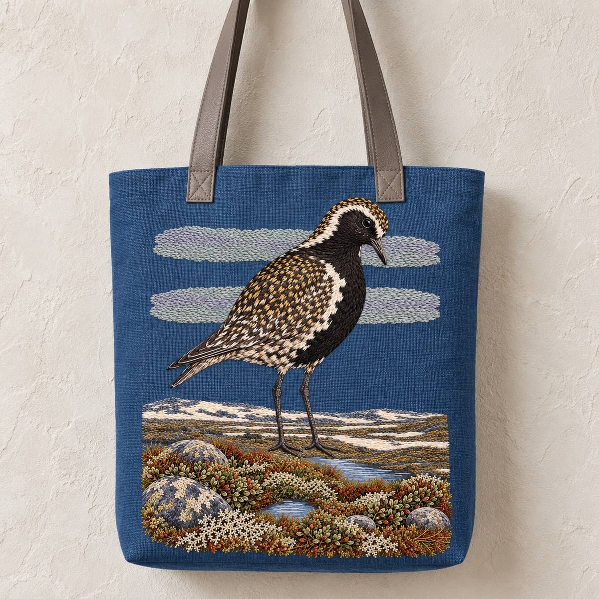 Golden Plover embroidery design in a digital mockup of A portrait project tote with long muted grey leather handles, storm-blue face and flat base, hanging against chalk plaster. Placement inspiration for Study 68.