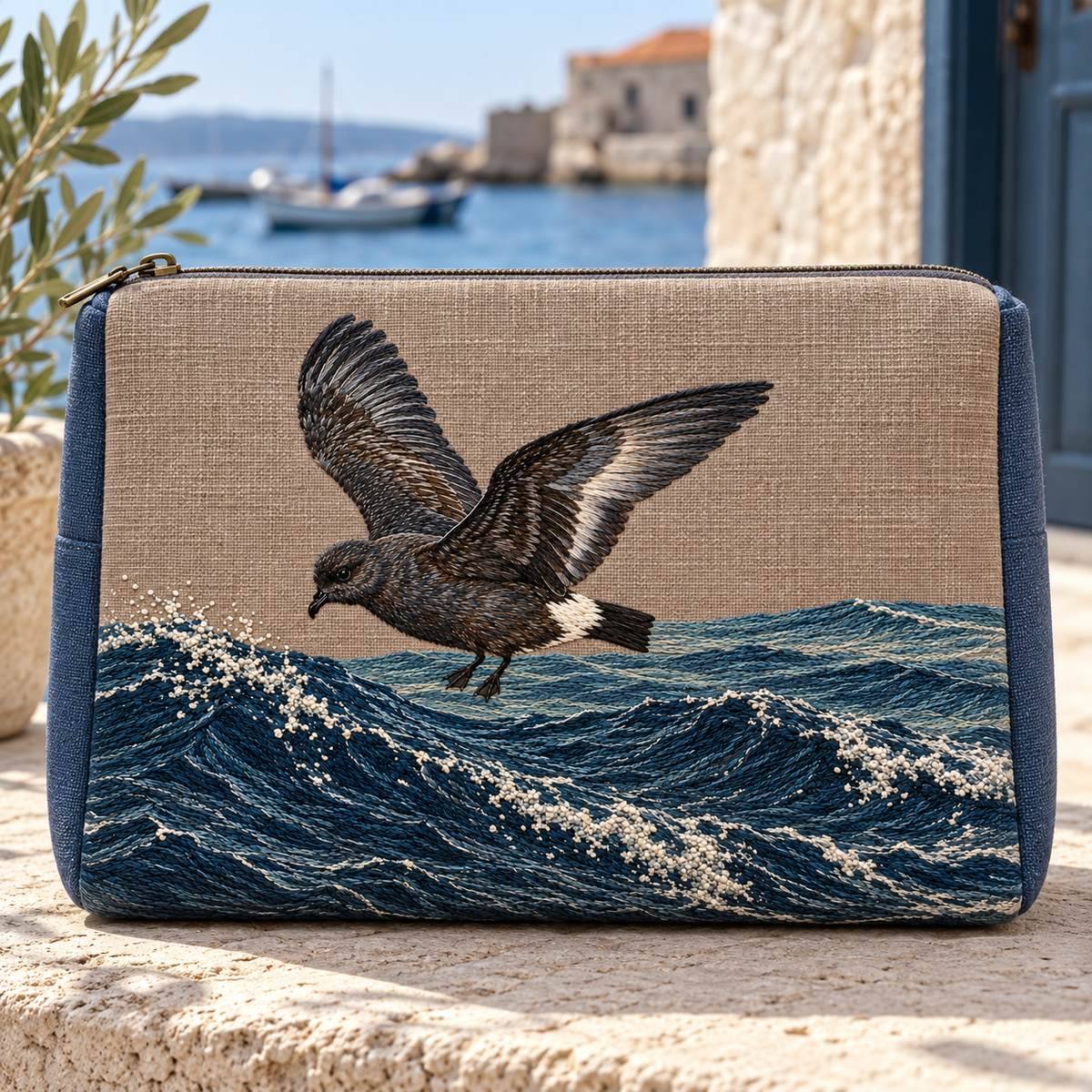Storm Petrel embroidery design in a digital mockup of A broad soft linen travelling case on a pale stone bench near a harbour doorway, copper-grey front, blue plain sides. Placement inspiration for Study 69.
