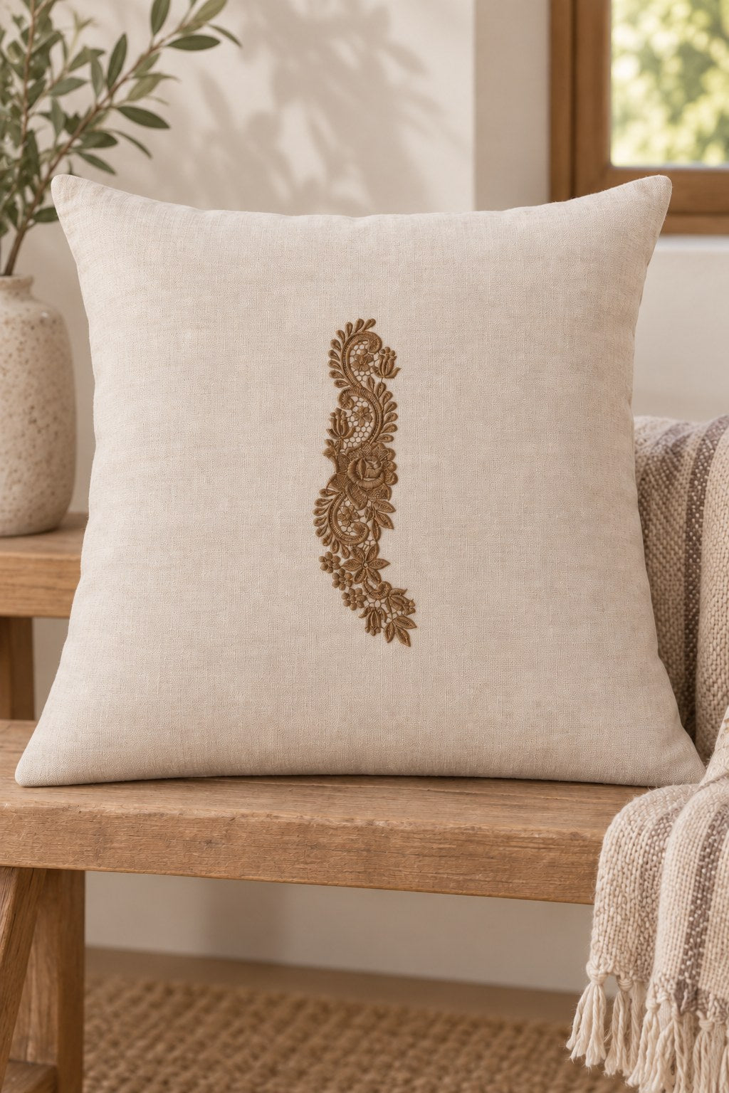 Styled linen cushion mockup with a single muted dark bronze brown rose and tulip lace spray embroidered on pale linen; the complete motif has clear fabric around it.