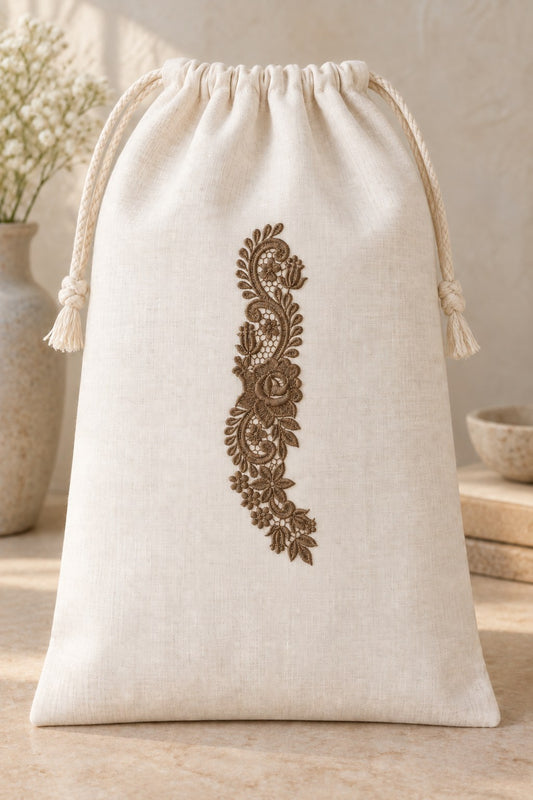 Styled linen drawstring bag mockup with a single muted dark bronze brown rose and tulip lace spray embroidered on pale linen; the complete motif has clear fabric around it.