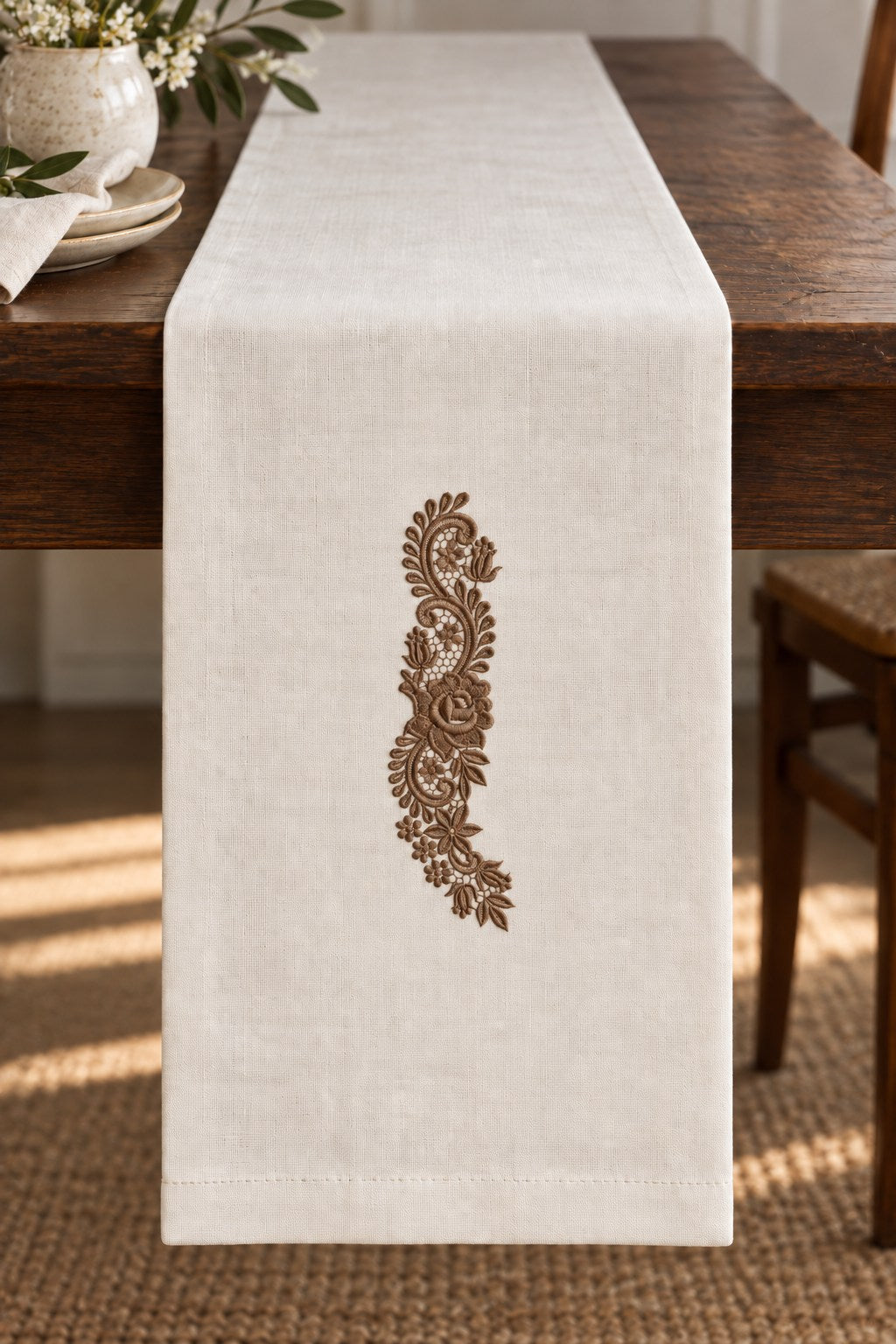 Styled linen table runner mockup with a single muted dark bronze brown rose and tulip lace spray embroidered on pale linen; the complete motif has clear fabric around it.