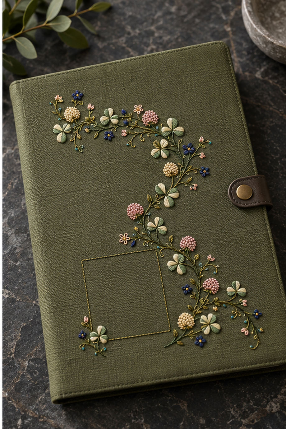 Green textile flat lay mockup featuring the Clover Ribbon Garland hand embroidery design. Clover, field blossoms and tendrils follow an S-shaped curve around a lower-left square.