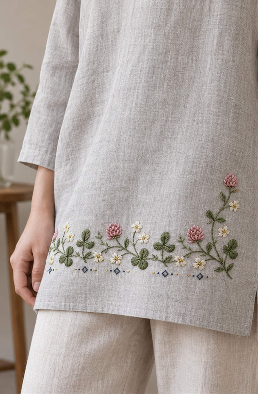 Linen garment hem mockup featuring the Clover Running Band hand embroidery design. Clover and daisies form a low horizontal band on white linen, ending with a taller right-hand stem.