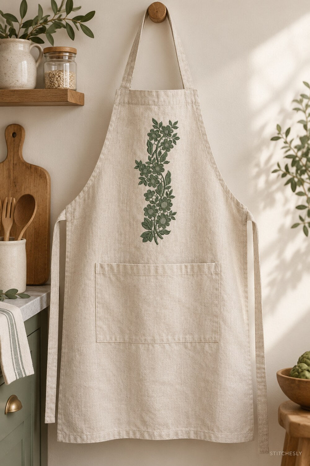 Generated usage mockup of a linen kitchen apron with the lattice blossom vine machine embroidery motif in one deep muted eucalyptus green thread color.