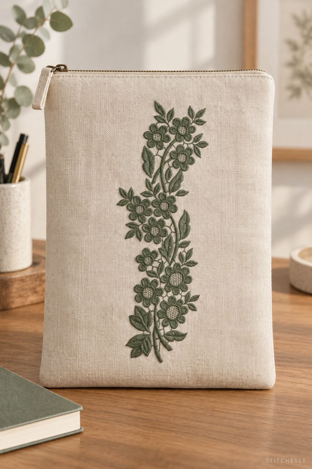 Generated usage mockup of a linen zip folio with the lattice blossom vine machine embroidery motif in one deep muted eucalyptus green thread color.