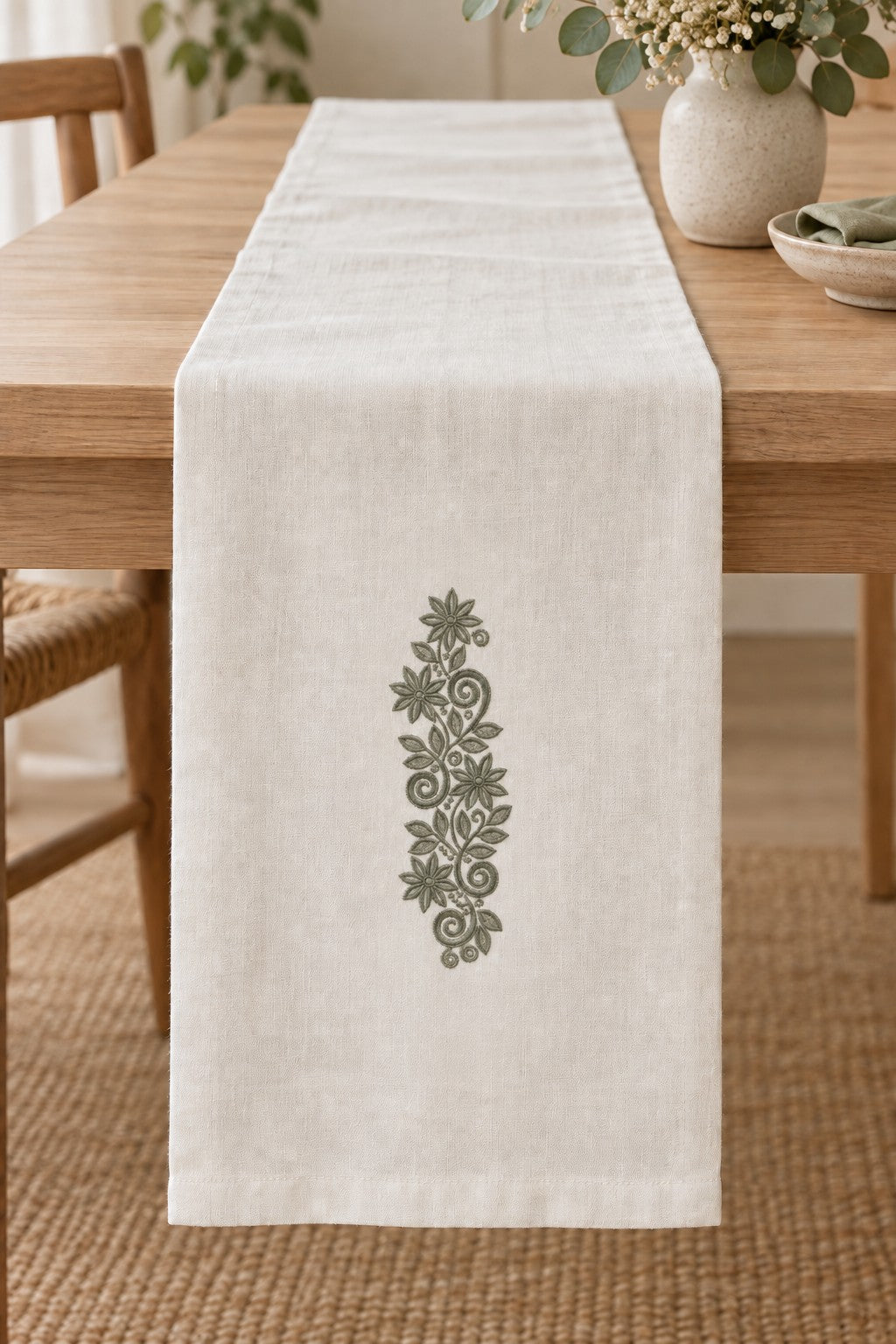 Styled linen table runner mockup with a single deep muted eucalyptus green starflower scroll vine embroidered on pale linen; the complete motif has clear fabric around it.