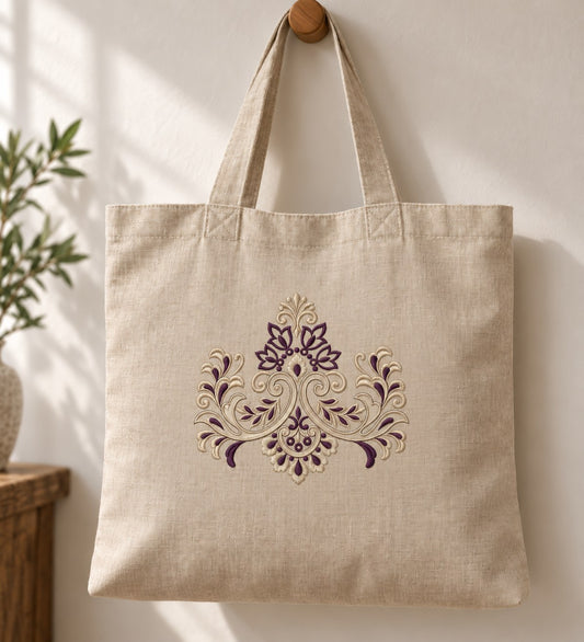 Symmetrical Floral Damask Crest embroidered in champagne and aubergine on a linen tote bag, showing the complete two-color motif.