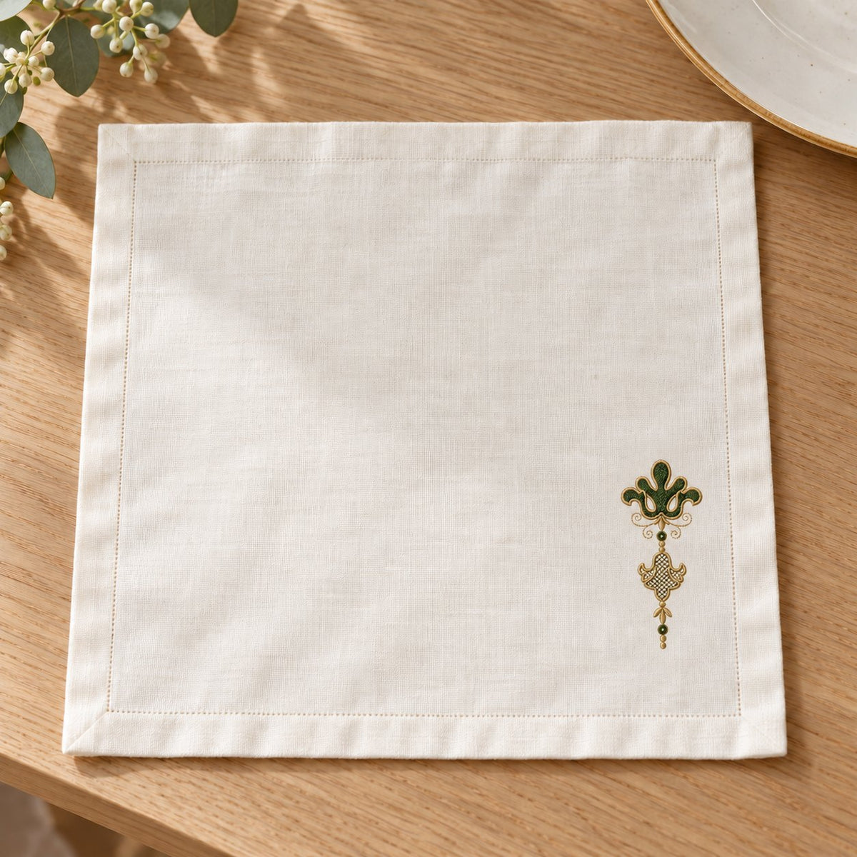 Linen napkin with a small green-and-champagne fleur de lis and lattice pendant near the lower corner.