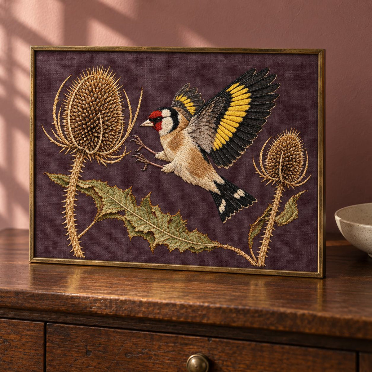 Goldfinch Across the Teasel Crown visual mockup shown in a frame leaning on a mantel, with the design on deep muted plum linen.