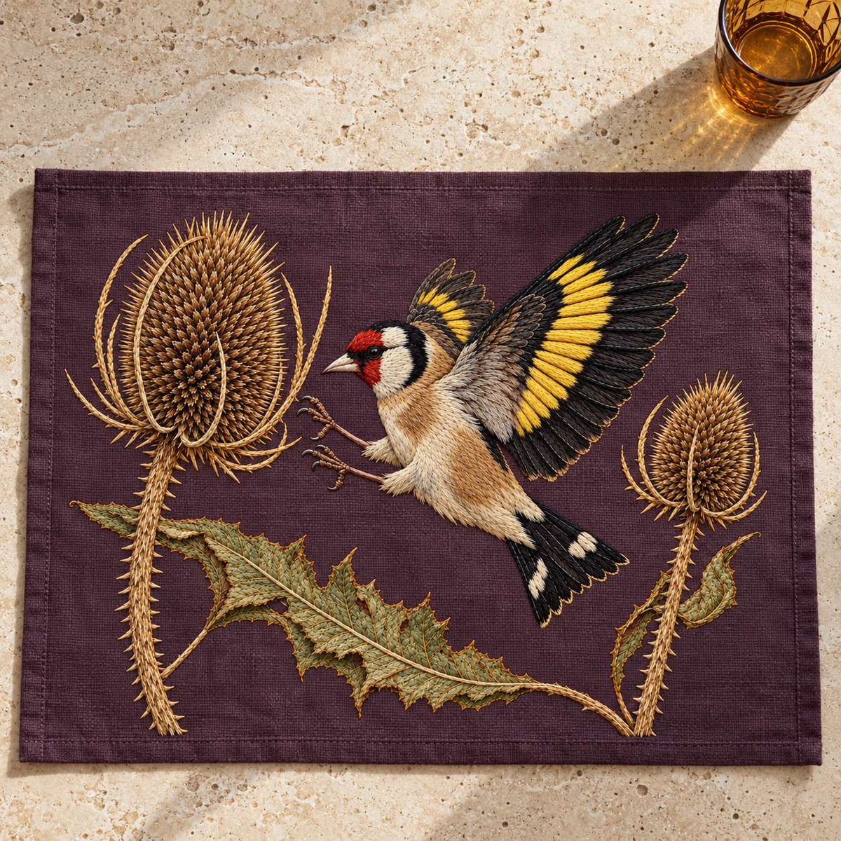 Goldfinch Across the Teasel Crown visual mockup shown as a textile panel on a stone platform, with the design on deep muted plum linen.