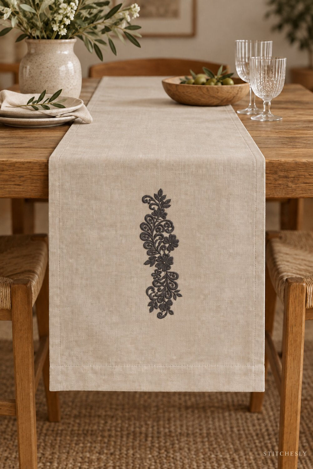 Generated usage mockup of a linen table runner with the teardrop leaf floral vine machine embroidery motif in one soft deep graphite charcoal thread color.