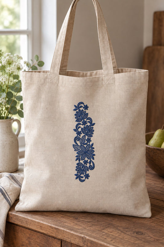 Styled linen market tote mockup with a single deep smoky indigo blue fan flower botanical spray embroidered on pale linen; the complete motif has clear fabric around it.